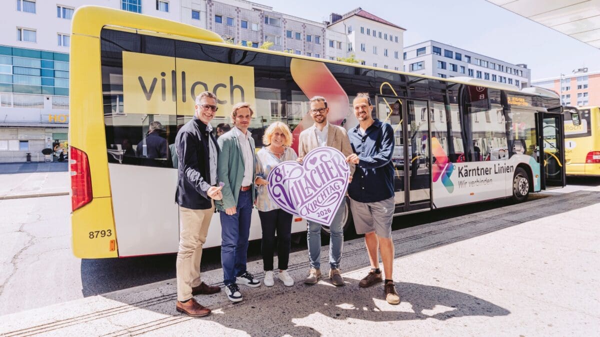 Vier Personen stehen vor einem gelben Bus und halten ein herzförmiges Schild für den Villacher Kirchtag.