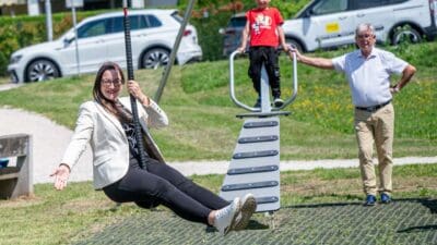 Eine Frau in weißem Blazer schwingt auf einer Affenschaukel, ein Junge steht auf der Konstruktion und ein Mann hält sie fest.