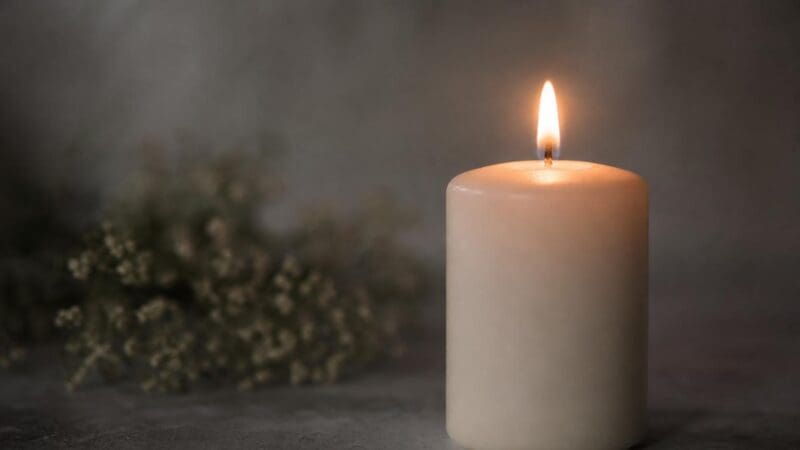 Eine brennende weiße Kerze mit heller Flamme und unscharfen getrockneten Blumen im Hintergrund.