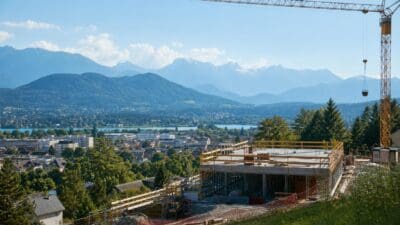 Betonrohbau mit gelbem Turmkran vor Stadt am See und Bergpanorama