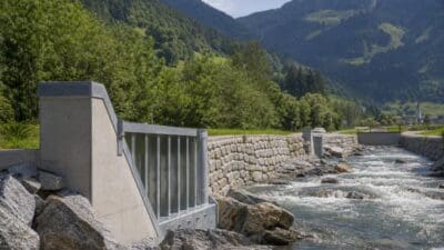 Betonbauwerk und Steinmauer am reißenden Gebirgsbach vor bewaldeten Bergen