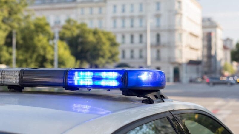 Blaues Blaulicht auf dem Dach eines Polizeifahrzeugs vor unscharfen Gebäuden und Bäumen.