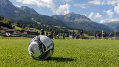 Fußball mit bunten Mustern liegt auf grünem Rasen vor Trainingsspielern in Tiroler Berglandschaft.
