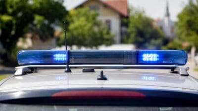 Blaue Blaulichter leuchten auf dem Dach eines silbernen Polizeifahrzeugs vor Wohnhäusern und Bäumen