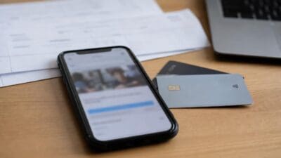 Smartphone mit unscharfem Display liegt neben Kreditkarten auf einem Holztisch mit Papieren und Laptop im Hintergrund