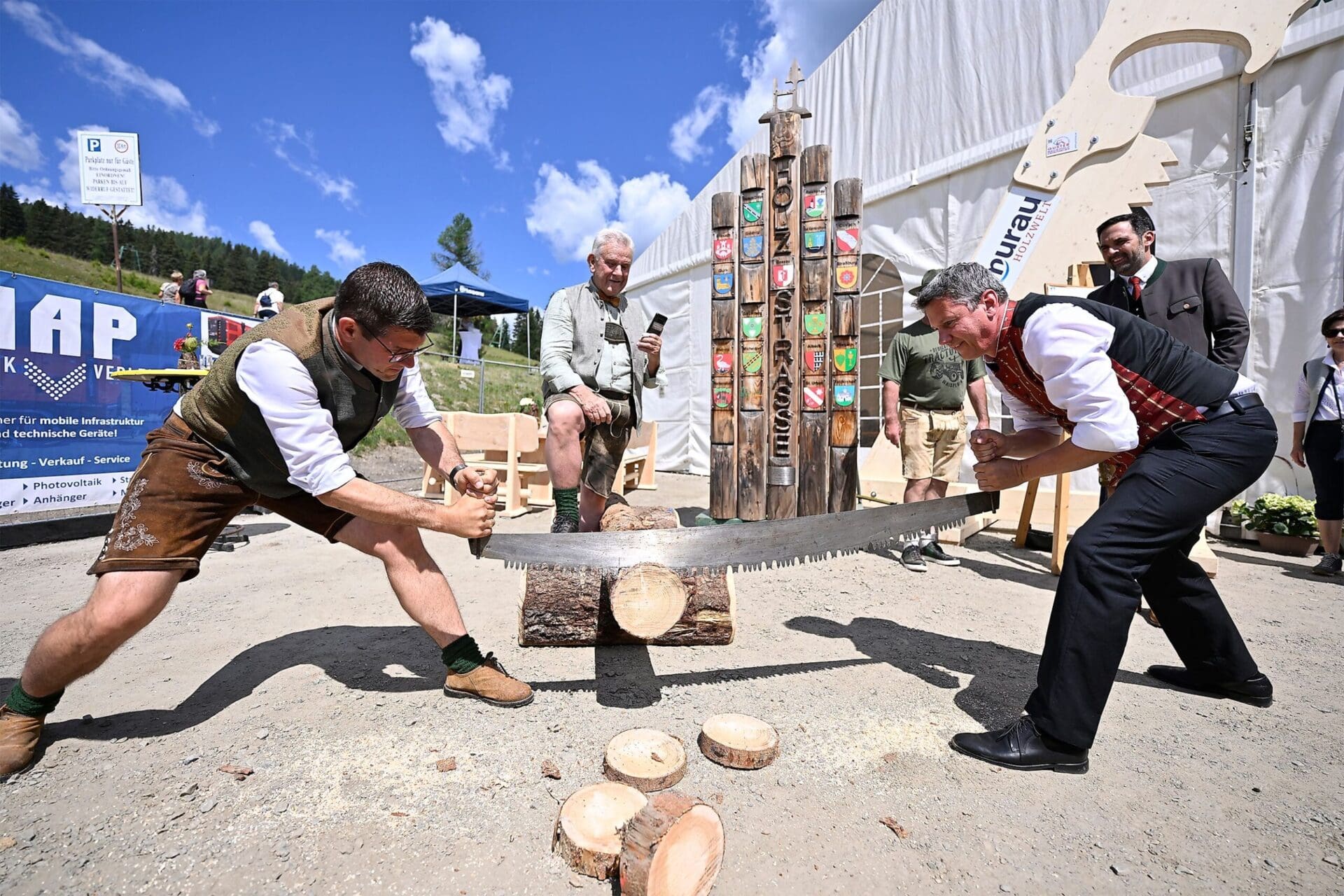 Zwei Männer in Tracht sägen mit einer großen Säge einen Holzstamm bei einem Fest.
