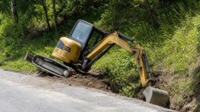 Ein gelber Bagger liegt nach einem Unfall seitlich geneigt an einer Böschung neben einer Landstraße.