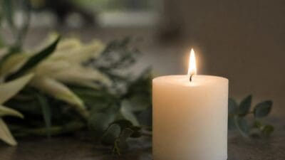 Eine brennende weiße Kerze steht vor weißen Blumen und grünen Blättern auf dunklem Untergrund.