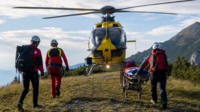 Rettungskräfte in roten Uniformen schieben eine Trage zu einem gelben Hubschrauber auf einem Bergweg.