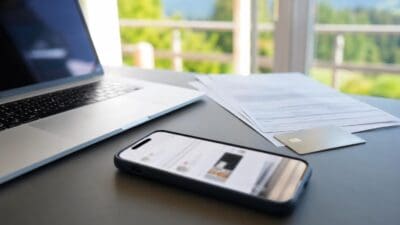 Ein silberner Laptop, ein Smartphone mit Chat-App, Dokumente und eine Bankkarte auf einem Schreibtisch.