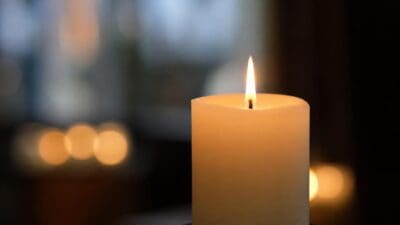 Eine brennende weiße Kerze mit heller Flamme vor unscharfem Hintergrund.