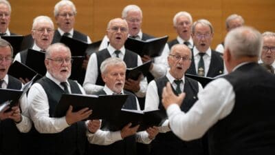 Ältere Männer in schwarzen Westen singen aus Notenheften unter Leitung eines Dirigenten