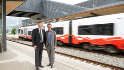 Zwei Männer in Anzügen stehen auf einem Bahnsteig vor einem vorbeifahrenden roten und weißen Zug.