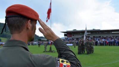 Soldat mit roter Baskenmütze salutiert vor der österreichischen Flagge auf einem Sportplatz
