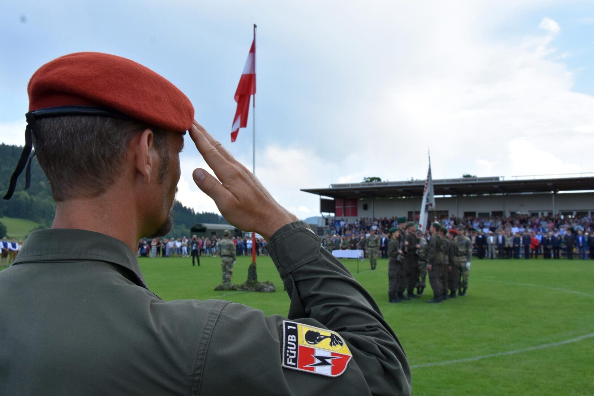 Soldat mit roter Baskenmütze salutiert vor der österreichischen Flagge auf einem Sportplatz