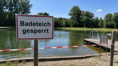 Weißes Schild mit roter Umrandung und Aufschrift Badeteich gesperrt vor einem See mit Holzsteg und Absperrband
