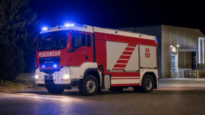 Rotes und weißes Feuerwehrfahrzeug mit aktiviertem Blaulicht vor einer Feuerwehrgarage bei Nacht
