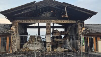 Ausgebranntes Feuerwehrhaus mit zerstörtem Dach und Trümmern hinter einem Metallzaun