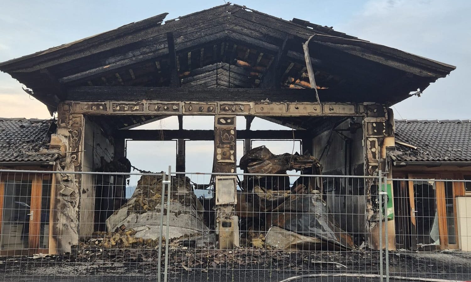 Ausgebranntes Feuerwehrhaus mit zerstörtem Dach und Trümmern hinter einem Metallzaun