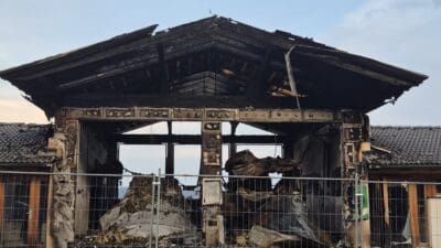 Ausgebranntes Feuerwehrhaus mit eingestürztem Dach und Trümmern hinter Absperrzaun