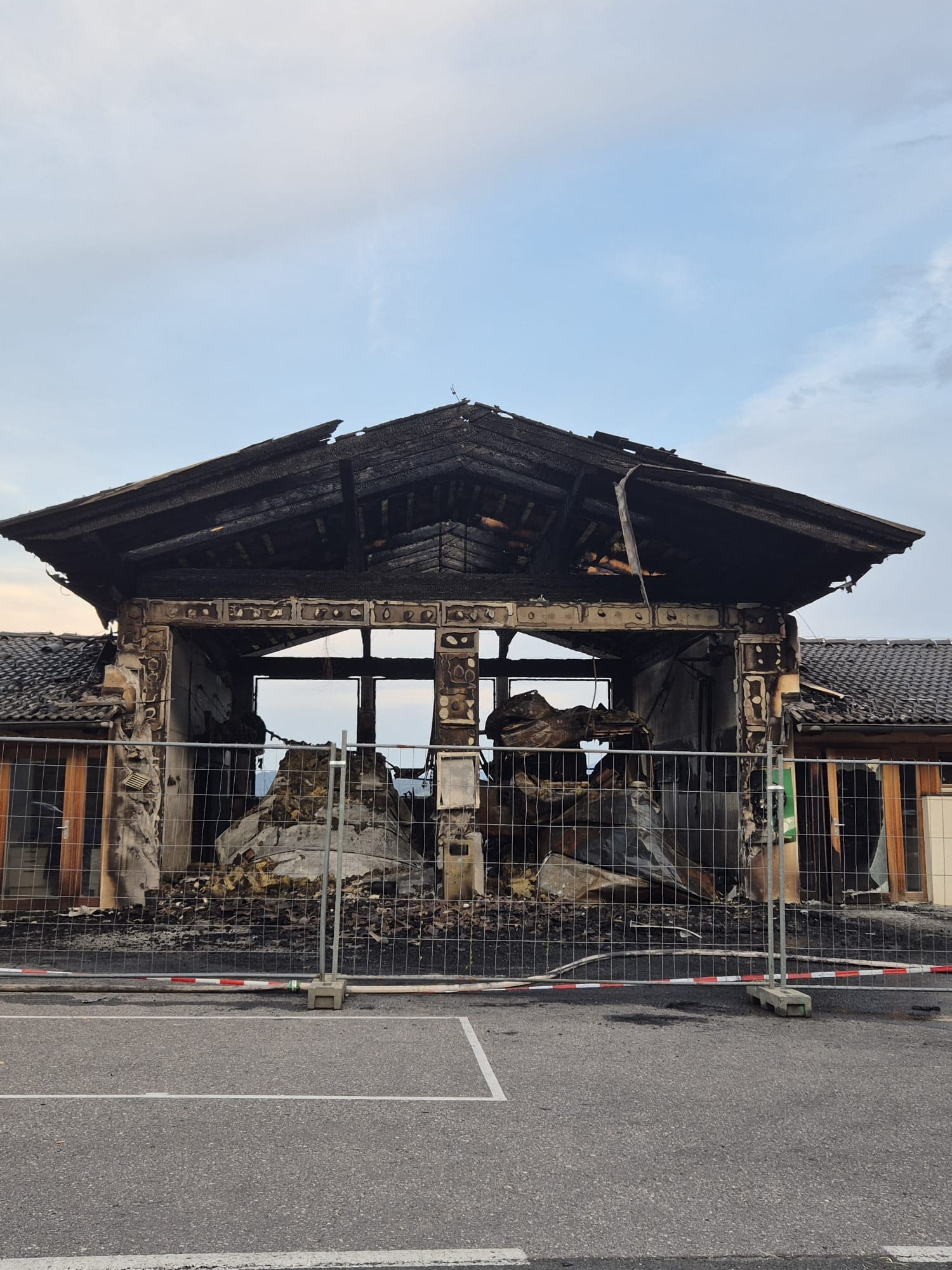 Ausgebranntes Feuerwehrhaus mit eingestürztem Dach und Trümmern hinter Absperrzaun