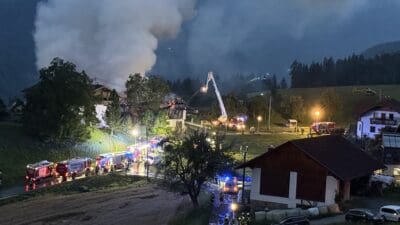 Nachtaufnahme eines brennenden Gebäudes mit starker Rauchentwicklung, umgeben von Feuerwehrfahrzeugen und Einsatzkräften.