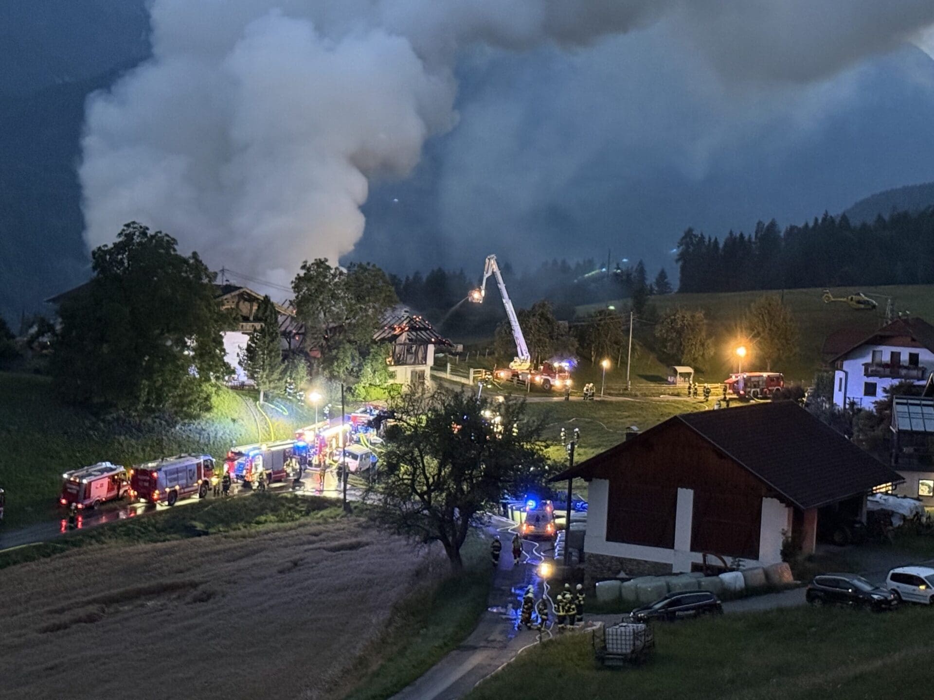 Nachtaufnahme eines brennenden Gebäudes mit starker Rauchentwicklung, umgeben von Feuerwehrfahrzeugen und Einsatzkräften.