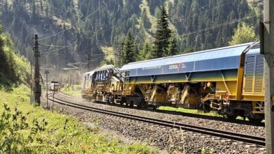 Ein gelb-blauer Gleisbauzug der Rhomberg Sersa Rail Group auf Bahngleisen in einem Tal.