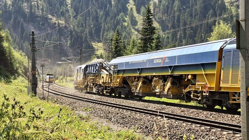 Ein gelb-blauer Gleisbauzug der Rhomberg Sersa Rail Group auf Bahngleisen in einem Tal.