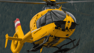 Gelber Rettungshubschrauber mit rot-weißem Heck fliegt über bewaldetem Gebirge