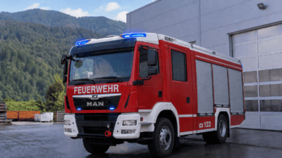 Rotes und weißes Feuerwehrfahrzeug MAN mit Blaulicht vor einer Halle und Bergen