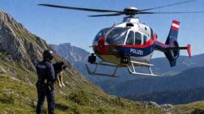 Polizist mit Hund steht vor schwebendem Polizeihubschrauber in Berglandschaft