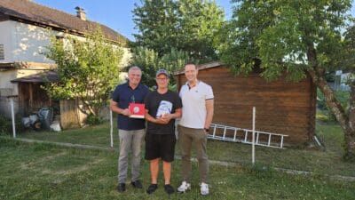 Drei Männer stehen nebeneinander auf einer Wiese vor einem Holzschuppen und lächeln.