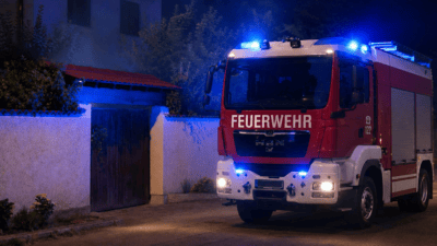 Rotes Feuerwehrfahrzeug mit leuchtenden blauen Blaulichtern in der Nacht vor einem Haus.