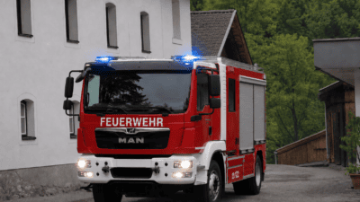 Rotes und weißes Feuerwehrfahrzeug mit Blaulicht vor einem Gebäude in ländlicher Umgebung
