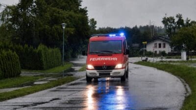 Rotes Feuerwehrauto mit blauem Blaulicht fährt auf nasser Straße durch eine ländliche Gegend.