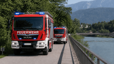 Zwei rote Feuerwehrfahrzeuge mit Blaulicht fahren auf einem Weg neben einem Fluss mit Bergen im Hintergrund.