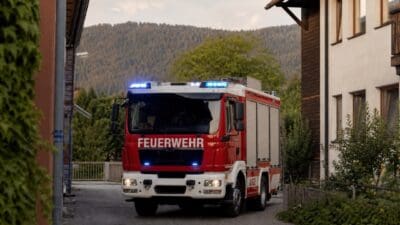Rotes und weißes Feuerwehrfahrzeug mit aktiviertem Blaulicht auf einer schmalen Dorfstraße.