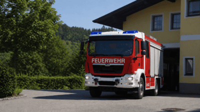 Rotes und weißes Feuerwehrfahrzeug mit Blaulicht vor gelber Feuerwache