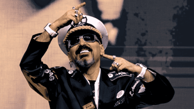 Gigi D’Agostino lächelt in weißer Kapitänsmütze und Sonnenbrille mit erhobenen Händen.