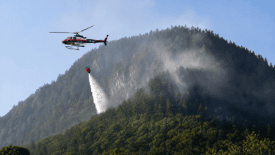 Ein Polizeihubschrauber lässt Wasser aus einem roten Behälter über einem bewaldeten Berg fallen