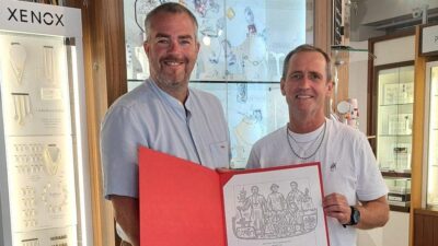 Zwei Männer präsentieren in einem Schmuckgeschäft ein geöffnetes Ehrendiplom in einem roten Ordner