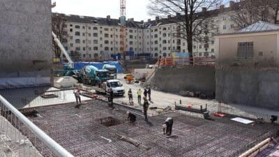 Bauarbeiter in Schutzkleidung auf einem großen Bewehrungsrost einer Baustelle mit Betonmischer und Kran