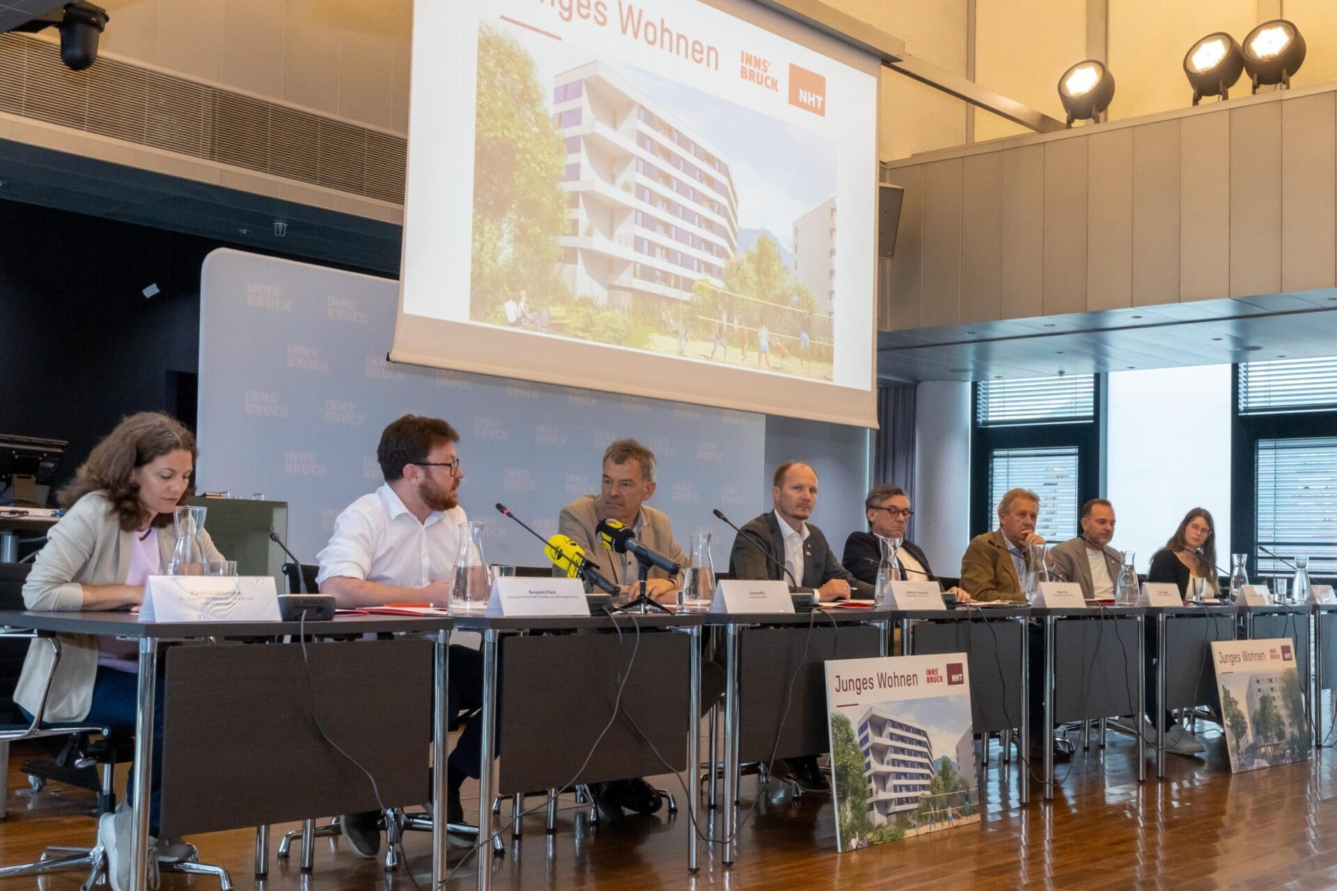 Mehrere Personen sitzen an einem Tisch während einer Pressekonferenz mit einer Projektion eines Wohngebäudes im Hintergrund