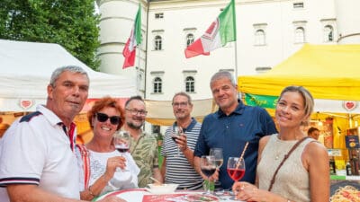 Fünf Personen mit Weingläsern an einem Tisch vor italienischen Flaggen und einem historischen Gebäude