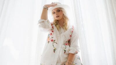Eine junge blonde Frau trägt einen weißen Cowboyhut und eine bestickte weiße Jacke vor Vorhängen.