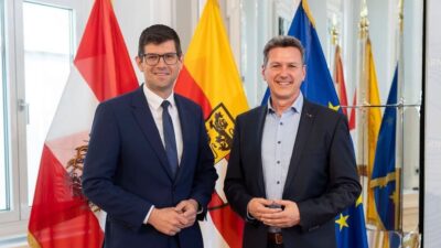 Zwei Männer in Anzügen stehen vor der österreichischen und Kärntner Flagge in einem hellen Saal