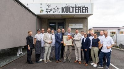 Mitarbeiter und Politiker posieren mit dem Kärntner Landeswappen vor dem Firmengebäude von Feine Küche Kulterer