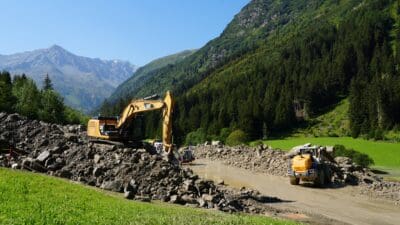 Gelber Bagger und Radlader räumen Steine und Schlamm von einer Straße in einem grünen Bergtal