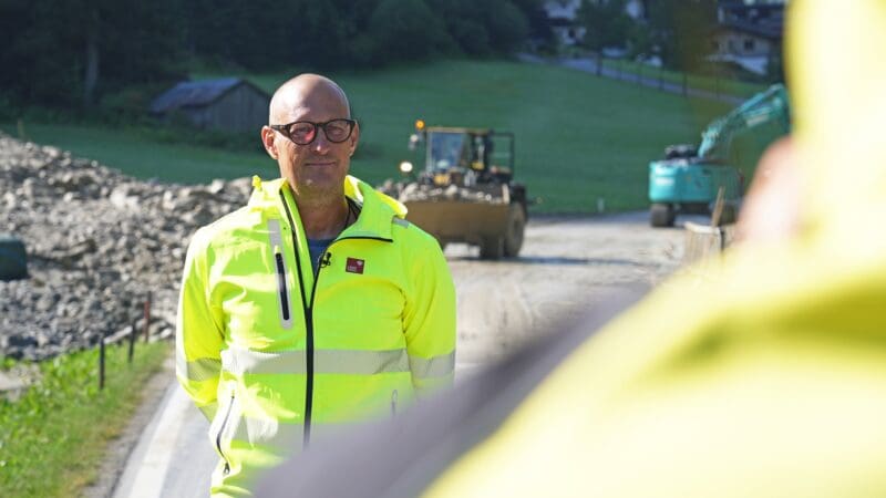 Ein Mann in neongelber Warnjacke steht vor Baggern auf einer Straße mit Geröll